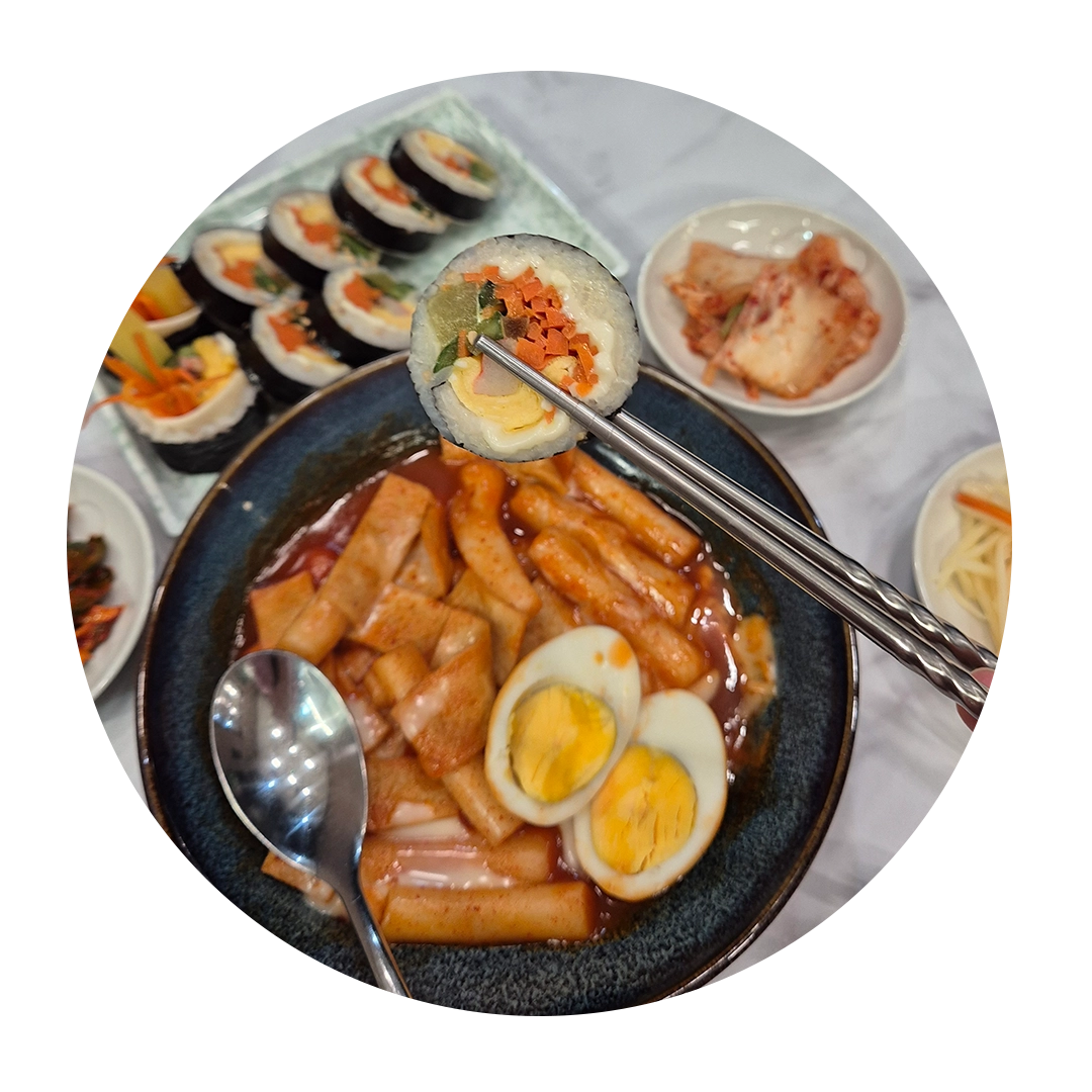 Chopsticks holding a piece of kimbap over a bowl of tteokbokki, with more kimbap and side dishes in the background.