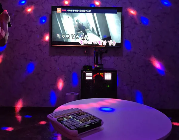 Karaoke room with disco lights, a television screen displaying a music video, and a control panel on a white table.