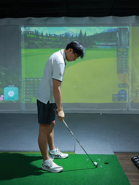 Golfer in a white polo shirt and shorts preparing to hit a ball on an indoor golf simulator.
